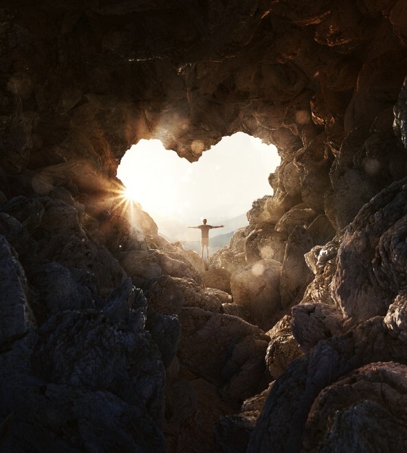 Mann steht mit ausgebreiteten Armen vor einer Herzförmigen Höhlenöffnung und blickt nach draußen